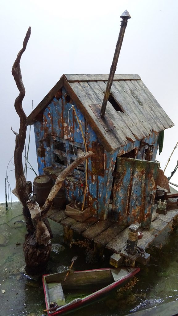Diorama artístico de una cabaña de madera azul envejecida sobre pilotes en un pantano, con efectos de agua estancada, un bote de remos y detalles de vegetación pantanosa.