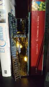 Book nook iluminado insertado entre libros en una estantería, con una callejuela medieval en miniatura con arcos de piedra, muros de ladrillo y pequeñas luces LED cálidas encendidas