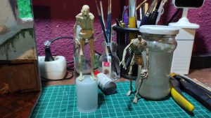Dos figuras esqueléticas humanoides en miniatura sobre la mesa de trabajo: una sin pintar en tono crudo y otra con acabado metálico oxidado, rodeadas de pinceles y materiales