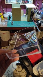 Caja de metacrilato transparente ensamblada a mano, sostenida frente a la mesa de trabajo del taller, junto a una estructura de DM en proceso de construcción al fondo