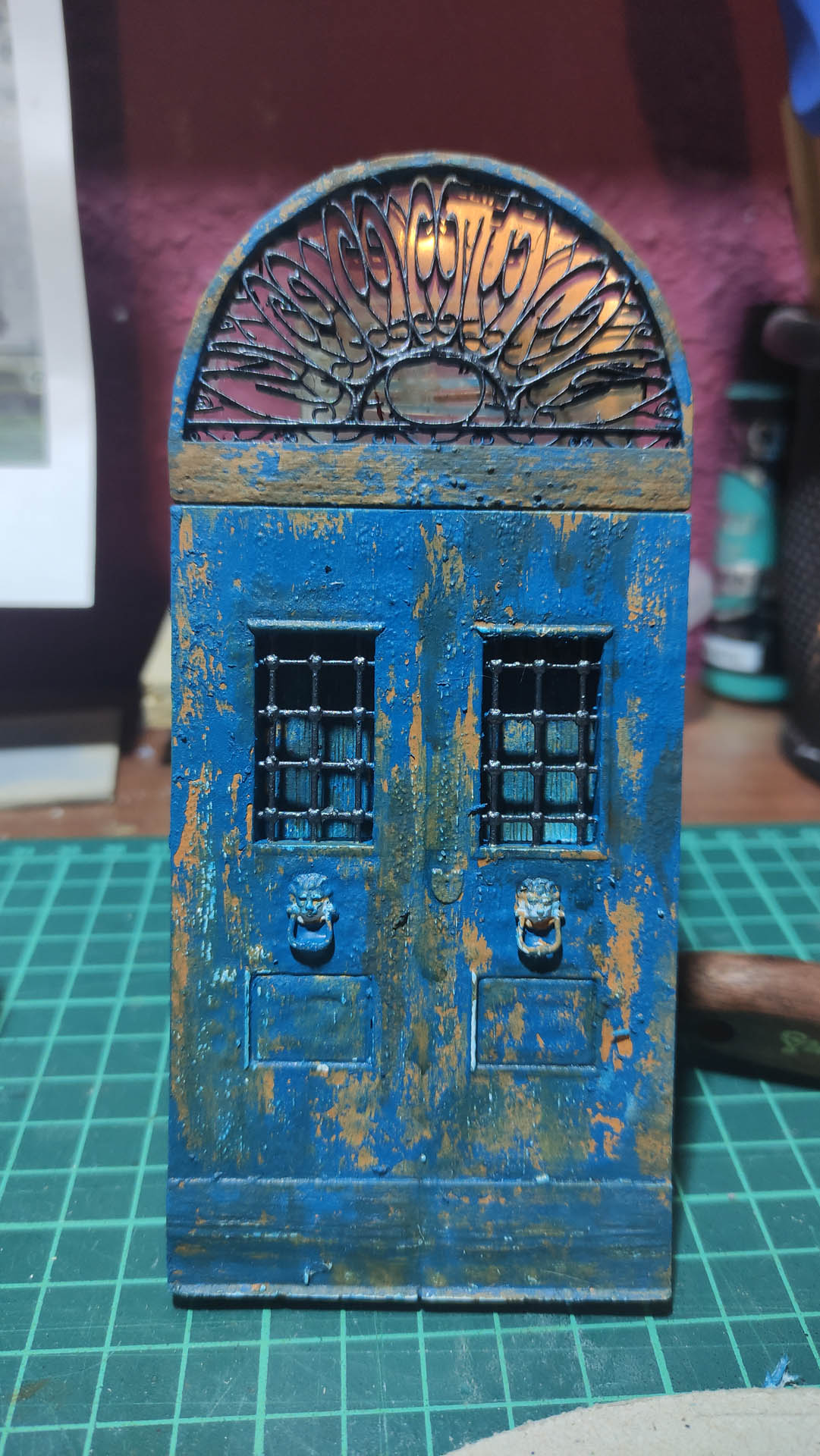 Puerta doble en miniatura de estilo victoriano envejecido, pintada en azul desconchado con acabado oxidado, con ventanillas enrejadas, aldabas con cabeza de león y lucernario semicircular con decoración calada