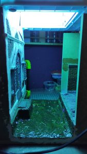 Proceso de curado de resina epoxi con luz ultravioleta en un diorama de Venecia en miniatura, donde el resplandor azul-verde de los LEDs UV ilumina el canal de agua simulado con resina aún en proceso de secado, junto a una fachada de edificio veneciano con ventana enrejada y muro de ladrillo