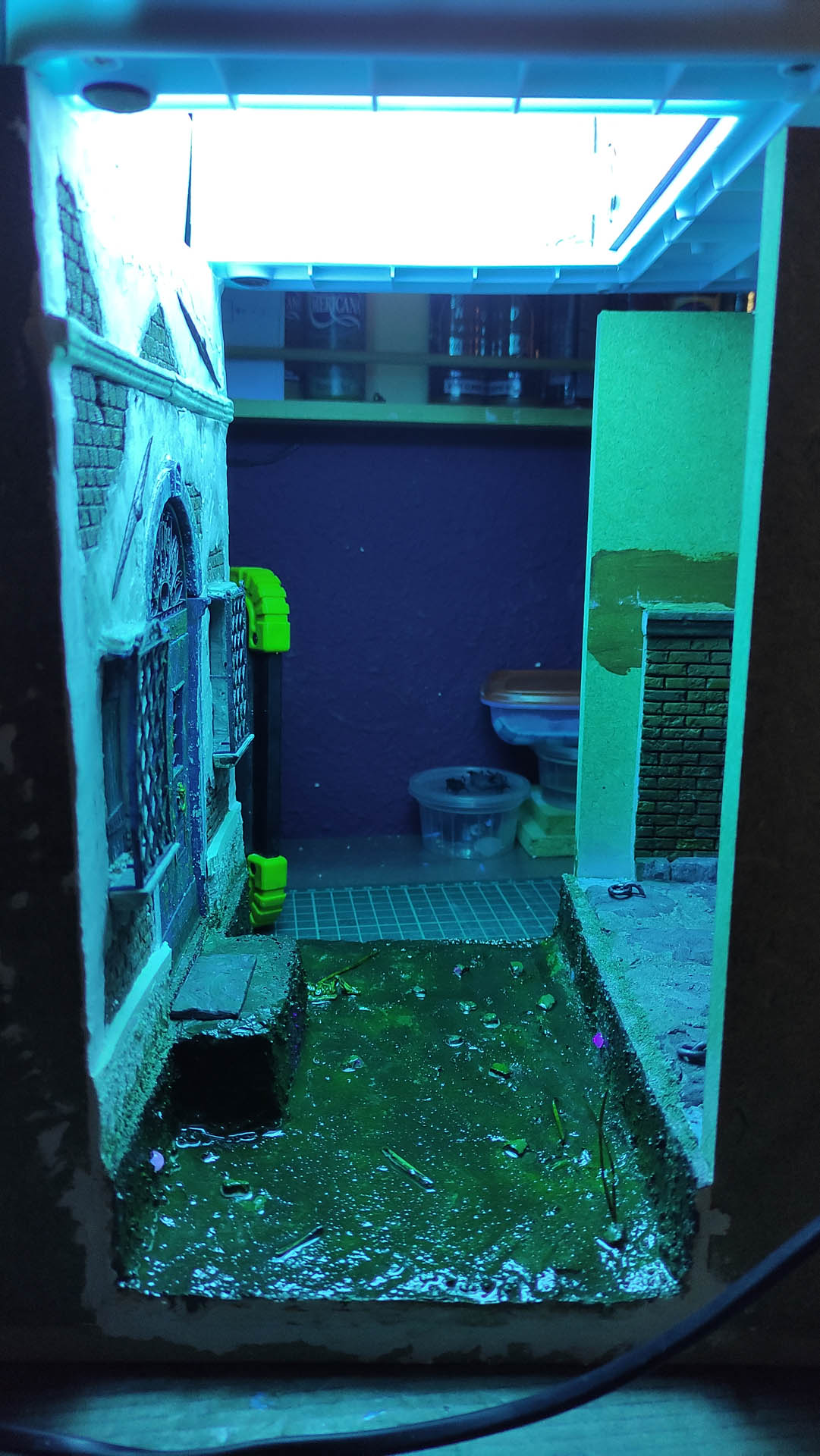 Proceso de curado de resina epoxi con luz ultravioleta en un diorama de Venecia en miniatura, donde el resplandor azul-verde de los LEDs UV ilumina el canal de agua simulado con resina aún en proceso de secado, junto a una fachada de edificio veneciano con ventana enrejada y muro de ladrillo