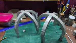 Dos estructuras de arcos entrelazados construidas con foam o cartón pintado de gris, simulando ruinas o elementos arquitectónicos góticos, sobre tapete de corte verde