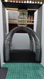 Arco de piedra en miniatura texturizado y pintado en gris, con sillares tallados uno a uno, colocado dentro de la estructura de un diorama en construcción