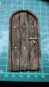 Puerta arqueada en miniatura elaborada con poliestireno extruido, tallado y texturizado para simular tablillas de madera envejecida con veta pronunciada y musgo, con cerrojo metálico en miniatura, apoyada sobre tapete de corte.