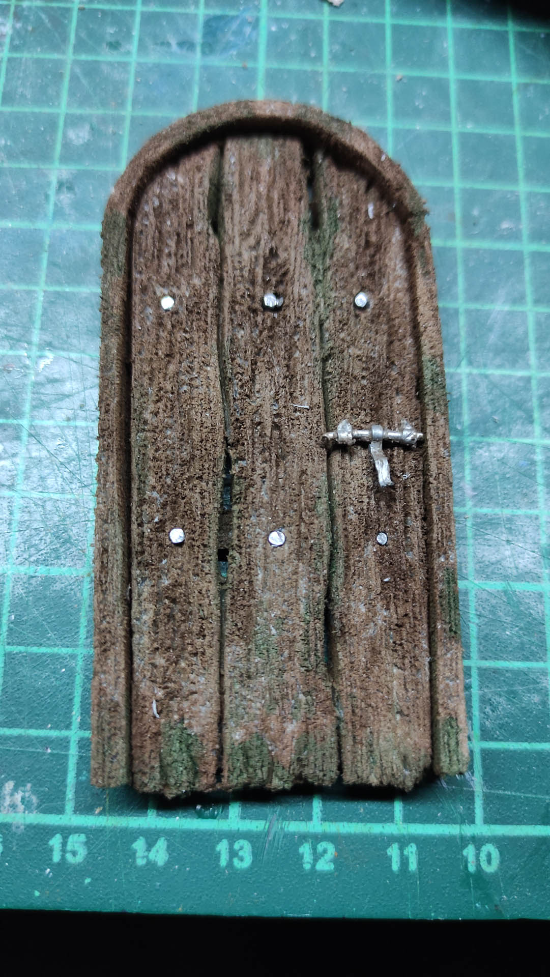 Puerta arqueada en miniatura elaborada con poliestireno extruido, tallado y texturizado para simular tablillas de madera envejecida con veta pronunciada y musgo, con cerrojo metálico en miniatura, apoyada sobre tapete de corte.