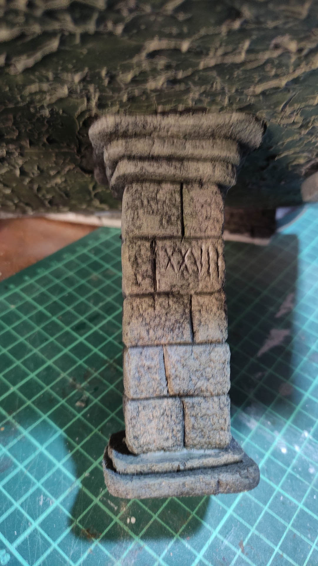 Columna de piedra en miniatura con capitel y basa, texturizada y pintada con técnica de lavado gris oscuro, con números romanos "XXVII" grabados en el fuste