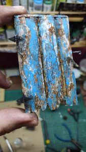 Fragmento de pared o valla en miniatura con tablillas de madera pintadas en azul desconchado y blanco envejecido, con clavos metálicos en miniatura, sostenida entre los dedos para mostrar el detalle.