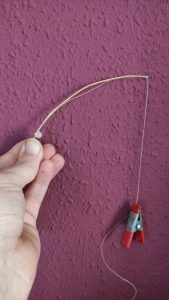 Mano sosteniendo una caña de pescar en miniatura hecha artesanalmente con una ramita curvada, hilo fino y una pinza roja metálica como contrapeso, ante una pared de estuco granate