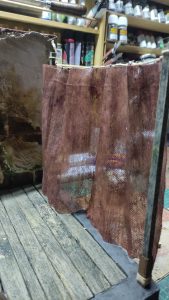 Cortina de tela de saco envejecida y manchada, colgada de una barra en miniatura sobre un suelo de tablones de madera, formando parte de un diorama de interior deteriorado.