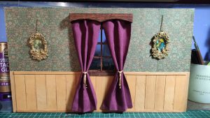 Pared de maqueta victoriana en miniatura con papel pintado verde floral, zócalo de madera, ventana con cortinas de tela morada recogidas con alzapaños dorados y dos pequeños cuadros con marcos barrocos dorados