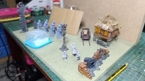 Vista general de las piezas del diorama de la Santa Compaña dispuestas sobre la mesa: figuras encapuchadas impresas en 3D sin pintar, una cruz procesional, figuritas de aldeanas, una cabaña con techo de paja, una carreta y elementos de paisaje rocoso.