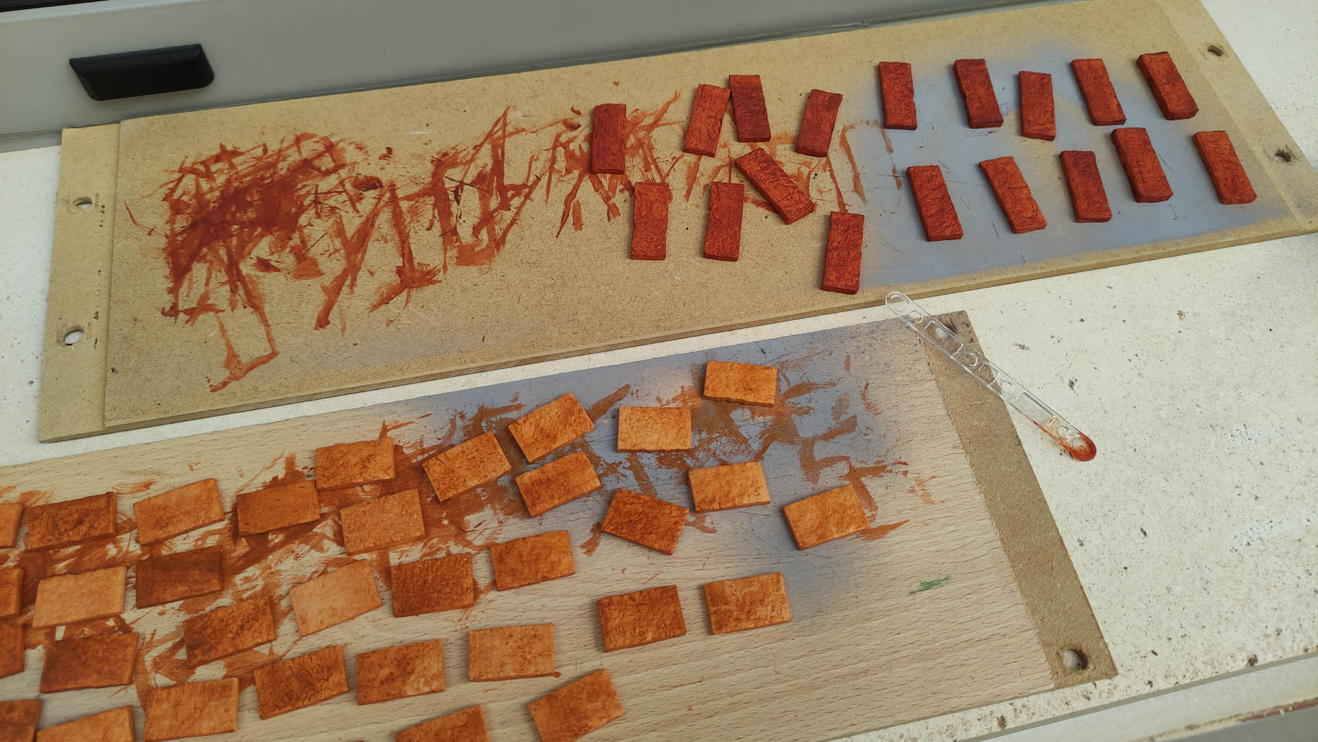 Proceso de pintado de ladrillos en miniatura: piezas rectangulares de foam o corcho en distintas fases de teñido, en tonos naranja y rojo óxido, sobre tablas de madera y superficie gris