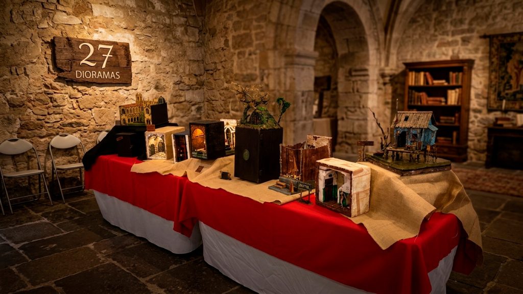 Vista general de la exposición de dioramas artesanales y maquetas en miniatura de 27 Dioramas en un salón de piedra rústica. La exhibición incluye una detallada catedral gótica, escenas interiores en cajas iluminadas, un diorama de bosque nocturno con vegetación natural preservada (A Santa Compaña) y una choza sobre pilotes en un pantano, todo dispuesto sobre mesas cubiertas con faldas rojas y arpillera. El fondo presenta estanterías llenas de libros antiguos y objetos de taller, y un cartel de madera de '27 DIORAMAS' colgado en la pared. La iluminación es cálida y enfocada, resaltando los detalles y efectos de luz de fibra óptica de cada obra