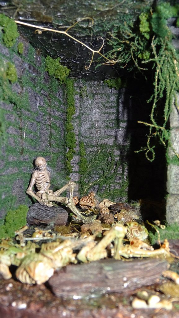 Miniatura artesanal de mazmorra medieval con esqueletos y vegetación de humedad. Diorama de realismo extremo sobre la Inquisición con texturas de piedra envejecida, hecho a mano por 27dioramas.