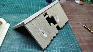 Diorama artístico de una cabaña de madera azul envejecida sobre pilotes en un pantano, con efectos de agua estancada, un bote de remos y detalles de vegetación pantanosa.