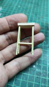 Diorama artístico de una cabaña de madera azul envejecida sobre pilotes en un pantano, con efectos de agua estancada, un bote de remos y detalles de vegetación pantanosa.