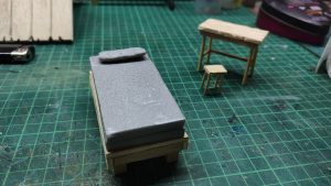 Diorama artístico de una cabaña de madera azul envejecida sobre pilotes en un pantano, con efectos de agua estancada, un bote de remos y detalles de vegetación pantanosa.