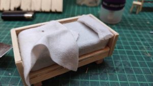 Diorama artístico de una cabaña de madera azul envejecida sobre pilotes en un pantano, con efectos de agua estancada, un bote de remos y detalles de vegetación pantanosa.