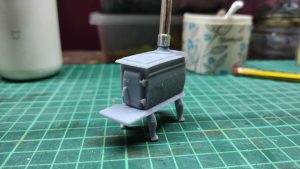 Diorama artístico de una cabaña de madera azul envejecida sobre pilotes en un pantano, con efectos de agua estancada, un bote de remos y detalles de vegetación pantanosa.