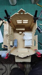 Diorama artístico de una cabaña de madera azul envejecida sobre pilotes en un pantano, con efectos de agua estancada, un bote de remos y detalles de vegetación pantanosa.