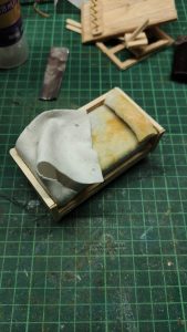 Diorama artístico de una cabaña de madera azul envejecida sobre pilotes en un pantano, con efectos de agua estancada, un bote de remos y detalles de vegetación pantanosa.