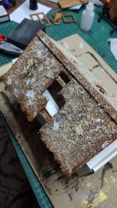 Diorama artístico de una cabaña de madera azul envejecida sobre pilotes en un pantano, con efectos de agua estancada, un bote de remos y detalles de vegetación pantanosa.