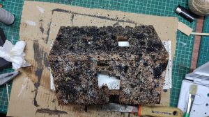 Diorama artístico de una cabaña de madera azul envejecida sobre pilotes en un pantano, con efectos de agua estancada, un bote de remos y detalles de vegetación pantanosa.