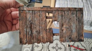 Diorama artístico de una cabaña de madera azul envejecida sobre pilotes en un pantano, con efectos de agua estancada, un bote de remos y detalles de vegetación pantanosa.