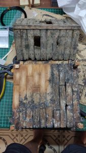 Diorama artístico de una cabaña de madera azul envejecida sobre pilotes en un pantano, con efectos de agua estancada, un bote de remos y detalles de vegetación pantanosa.