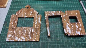 Diorama artístico de una cabaña de madera azul envejecida sobre pilotes en un pantano, con efectos de agua estancada, un bote de remos y detalles de vegetación pantanosa.