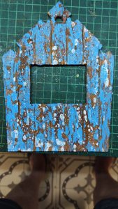 Diorama artístico de una cabaña de madera azul envejecida sobre pilotes en un pantano, con efectos de agua estancada, un bote de remos y detalles de vegetación pantanosa.