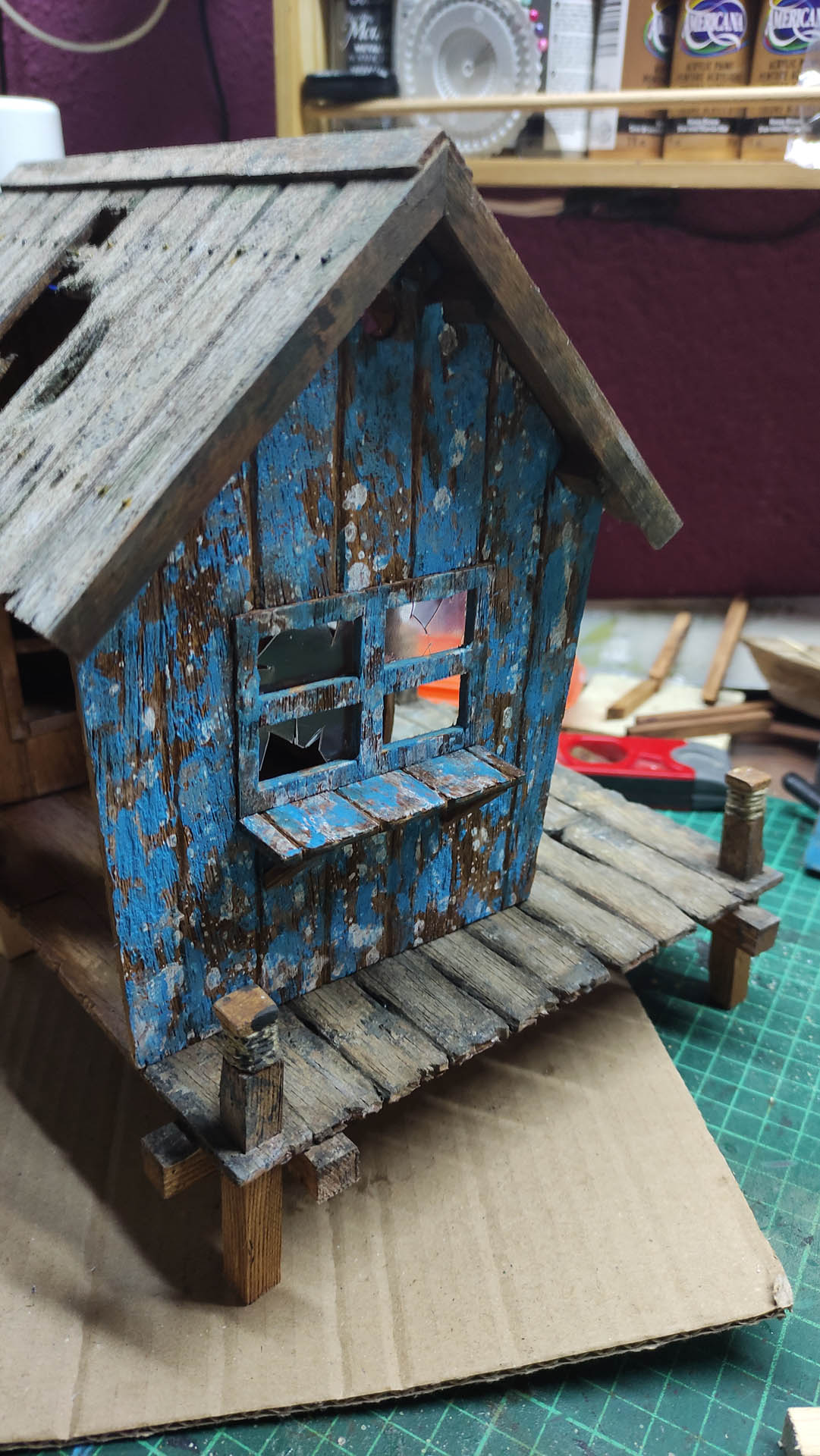 Diorama artístico de una cabaña de madera azul envejecida sobre pilotes en un pantano, con efectos de agua estancada, un bote de remos y detalles de vegetación pantanosa.