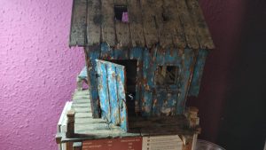 Diorama artístico de una cabaña de madera azul envejecida sobre pilotes en un pantano, con efectos de agua estancada, un bote de remos y detalles de vegetación pantanosa.