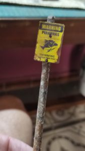 Diorama artístico de una cabaña de madera azul envejecida sobre pilotes en un pantano, con efectos de agua estancada, un bote de remos y detalles de vegetación pantanosa.