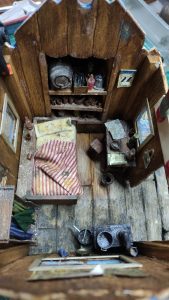 Diorama artístico de una cabaña de madera azul envejecida sobre pilotes en un pantano, con efectos de agua estancada, un bote de remos y detalles de vegetación pantanosa.