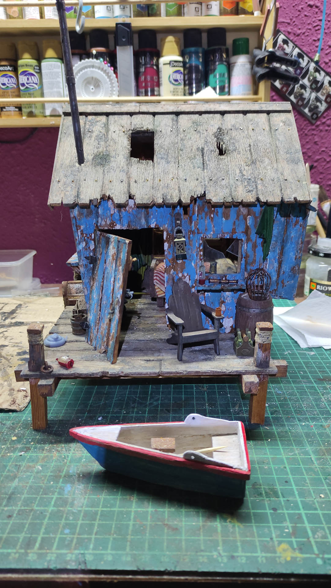 Diorama artístico de una cabaña de madera azul envejecida sobre pilotes en un pantano, con efectos de agua estancada, un bote de remos y detalles de vegetación pantanosa.