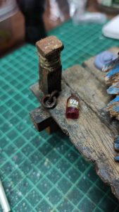 Diorama artístico de una cabaña de madera azul envejecida sobre pilotes en un pantano, con efectos de agua estancada, un bote de remos y detalles de vegetación pantanosa.