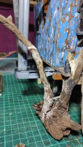 Diorama artístico de una cabaña de madera azul envejecida sobre pilotes en un pantano, con efectos de agua estancada, un bote de remos y detalles de vegetación pantanosa.