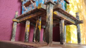 Diorama artístico de una cabaña de madera azul envejecida sobre pilotes en un pantano, con efectos de agua estancada, un bote de remos y detalles de vegetación pantanosa.