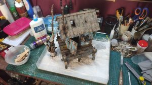 Diorama artístico de una cabaña de madera azul envejecida sobre pilotes en un pantano, con efectos de agua estancada, un bote de remos y detalles de vegetación pantanosa.