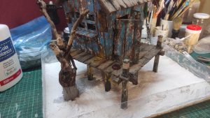 Diorama artístico de una cabaña de madera azul envejecida sobre pilotes en un pantano, con efectos de agua estancada, un bote de remos y detalles de vegetación pantanosa.