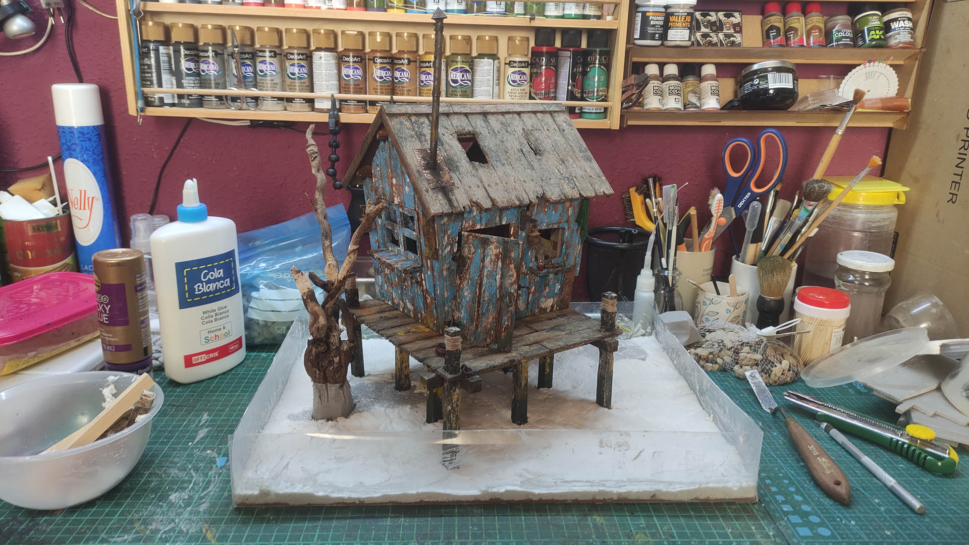 Diorama artístico de una cabaña de madera azul envejecida sobre pilotes en un pantano, con efectos de agua estancada, un bote de remos y detalles de vegetación pantanosa.