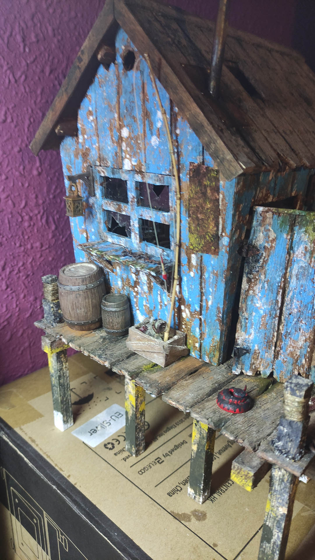 Diorama artístico de una cabaña de madera azul envejecida sobre pilotes en un pantano, con efectos de agua estancada, un bote de remos y detalles de vegetación pantanosa.