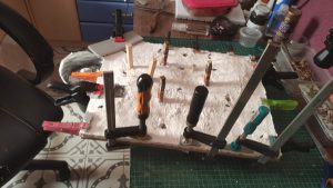 Diorama artístico de una cabaña de madera azul envejecida sobre pilotes en un pantano, con efectos de agua estancada, un bote de remos y detalles de vegetación pantanosa.