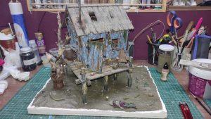 Diorama artístico de una cabaña de madera azul envejecida sobre pilotes en un pantano, con efectos de agua estancada, un bote de remos y detalles de vegetación pantanosa.