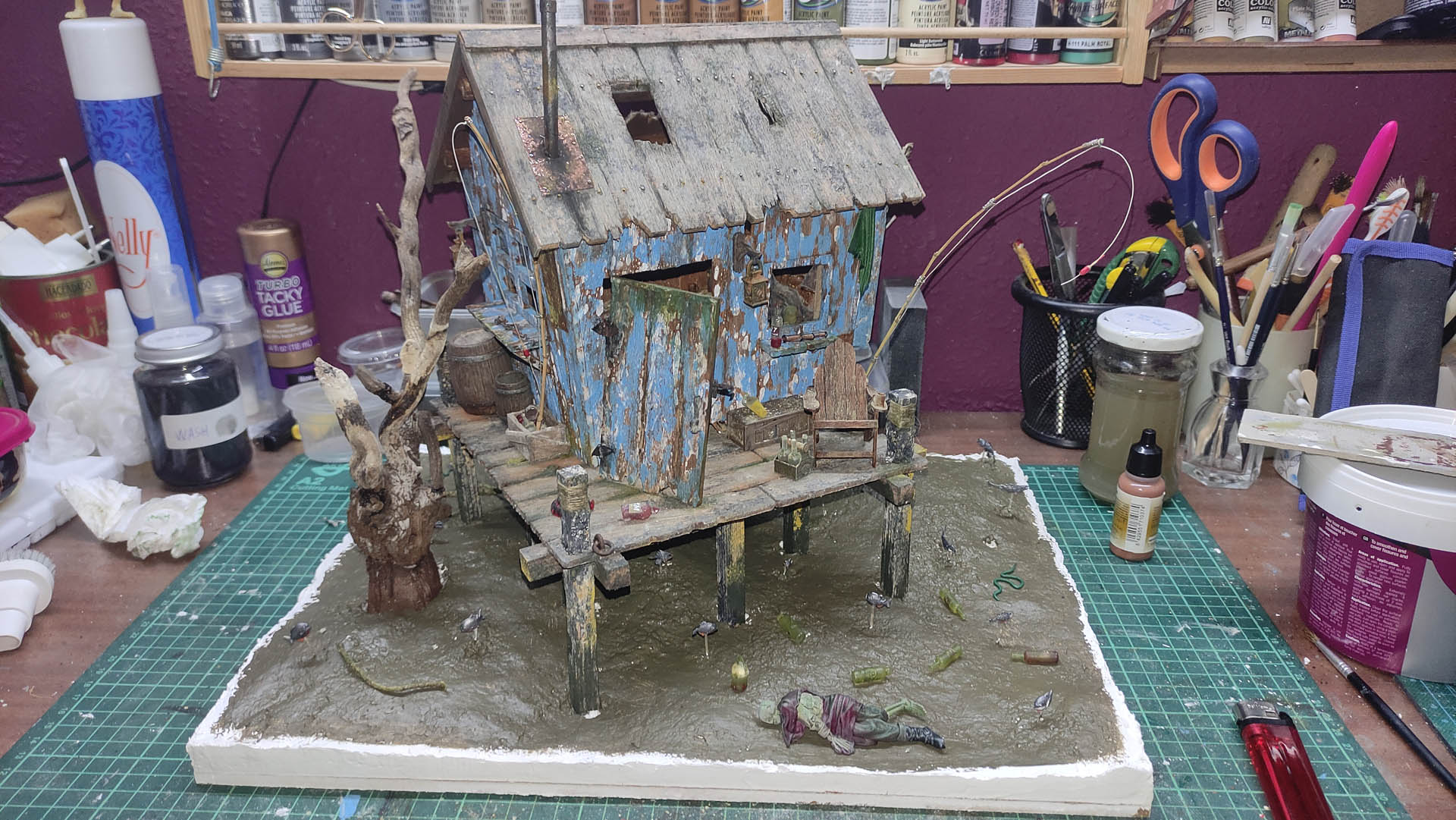 Diorama artístico de una cabaña de madera azul envejecida sobre pilotes en un pantano, con efectos de agua estancada, un bote de remos y detalles de vegetación pantanosa.