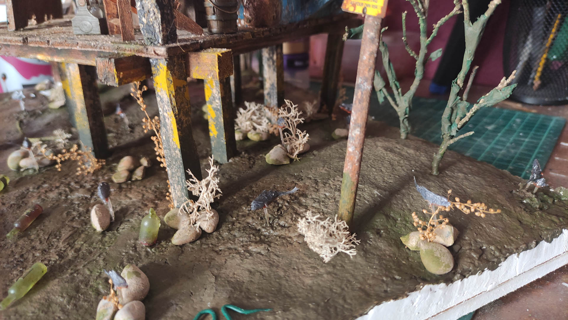 Diorama artístico de una cabaña de madera azul envejecida sobre pilotes en un pantano, con efectos de agua estancada, un bote de remos y detalles de vegetación pantanosa.