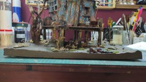 Diorama artístico de una cabaña de madera azul envejecida sobre pilotes en un pantano, con efectos de agua estancada, un bote de remos y detalles de vegetación pantanosa.